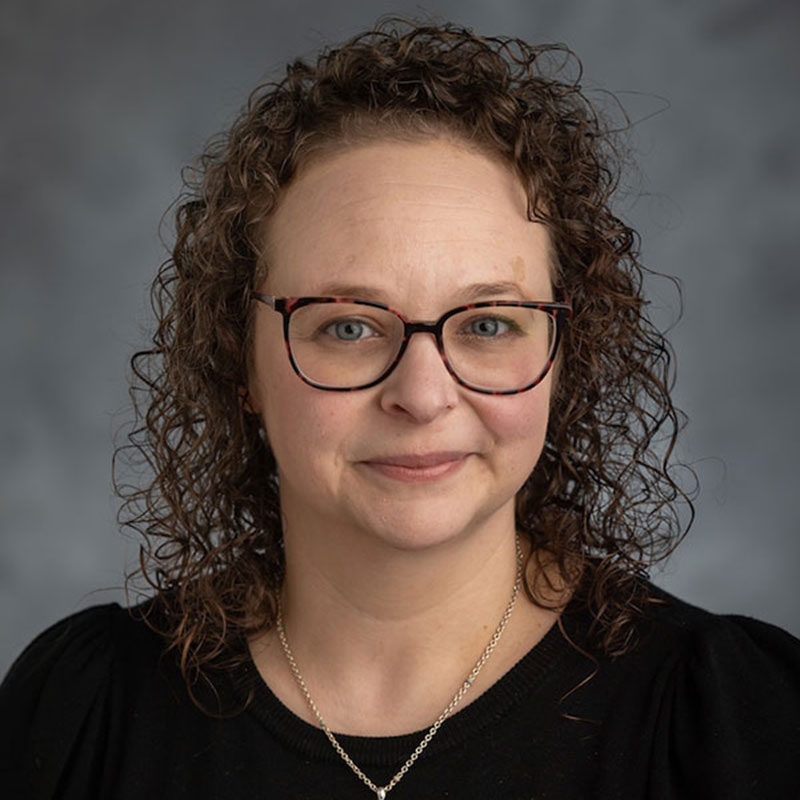 A woman with curly brown hair and glassed wearing a black blouse and necklace.
