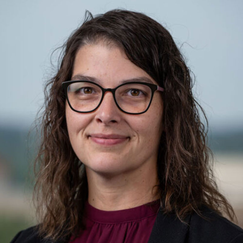 A woman with dark curly shoulder length hair and glasses wearing a maroon shirt and a dark jacket.