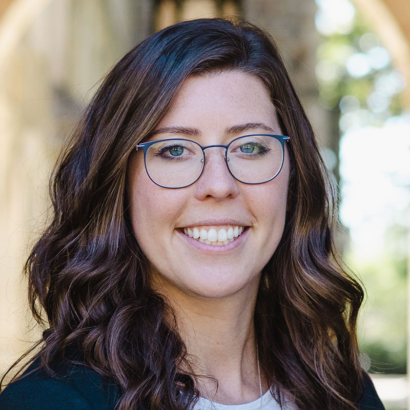 A woman with glasses and long curly brown hair wearing a white shirt and dark jacket.