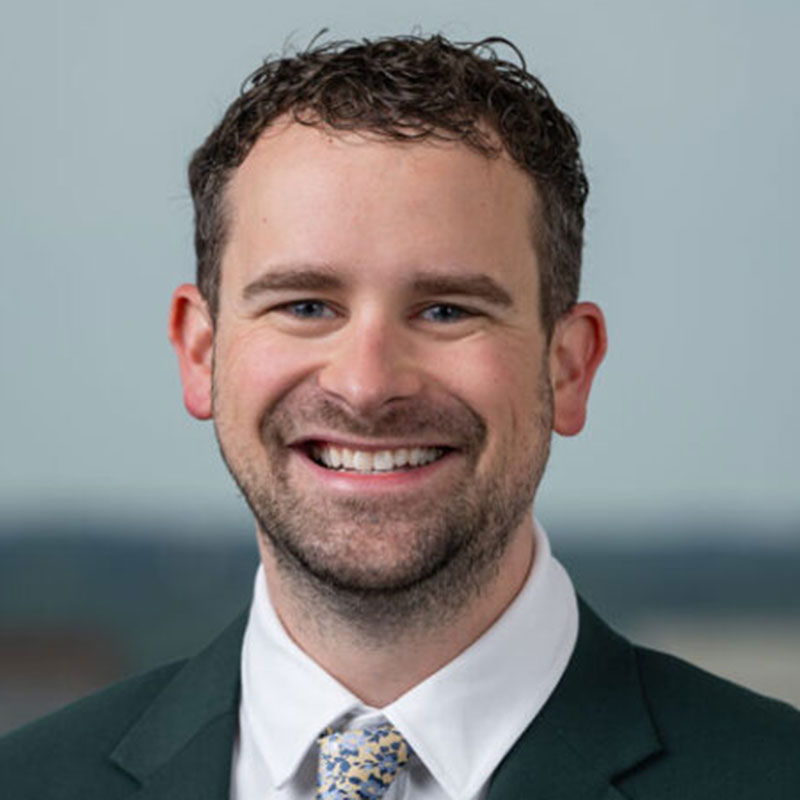 A man with short dark hair and beard stubble wearing a green suit with a tie.