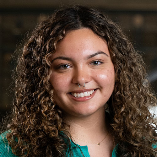 A woman with long, dark curly hair and a septum piercing wearing a teal sweater.