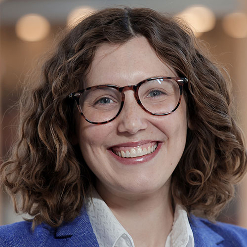 White woman with wavy auburn hair and tortoise rimmed glasses wearing a blue blazer.