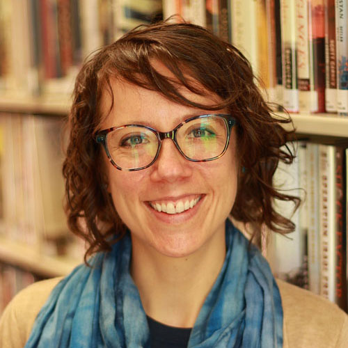 Woman with wavy shoulder length auburn hair and glasses with a blue scarf.