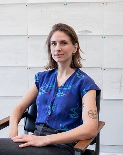 A white woman with chin length hair sits in a chair looking at the camera. She has a blue short sleeved shirt on and dark pants. Behind her are many drawings pinned to a wall. 