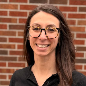 A white woman with long brown hair and glasses wearing a dark blouse.