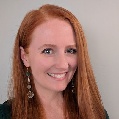 A white woman with long red hair wearing long earrings and a green blouse.