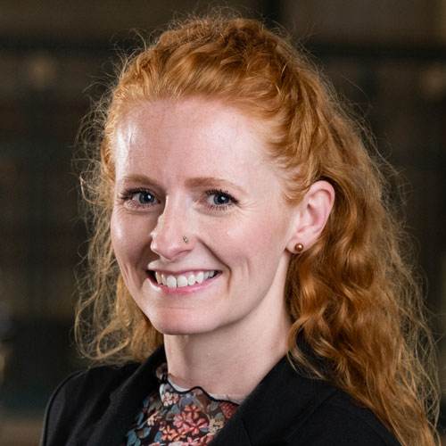 Woman with long curly red hair waring a bright pattern shirt and dark suit jacket.