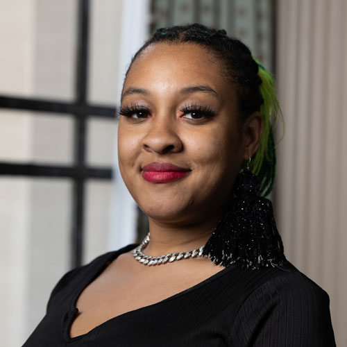 A woman with long dark hair with a green streak pulled back in box braids wearing a black blouse.